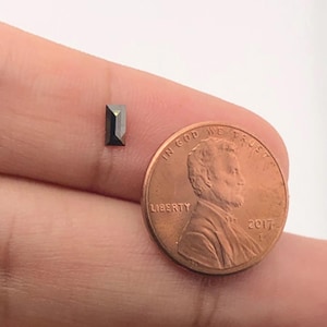 Puede incluir: Una pequeña piedra preciosa rectangular negra se sostiene en una mano junto a un centavo estadounidense para comparar el tamaño. "In God We Trust" y "Liberty" son visibles en el centavo.