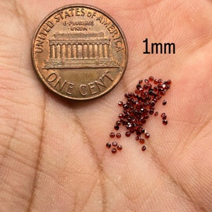 May include: A pile of small, dark red, 1 mm round gemstones next to a US penny for size comparison.  "1mm" is written next to the gemstones.