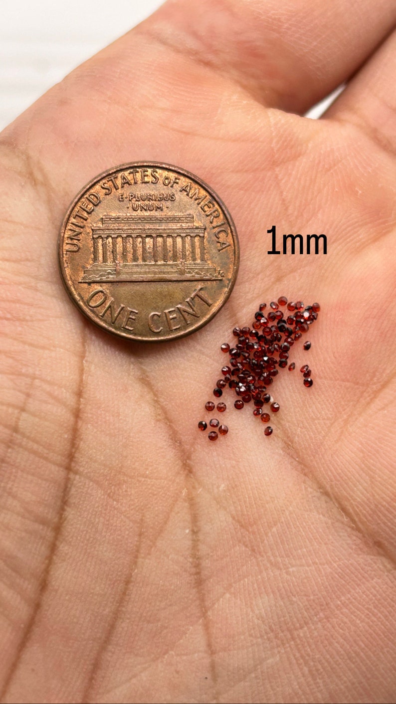 May include: A pile of small, dark red, 1 mm round gemstones next to a US penny for size comparison.  "1mm" is written next to the gemstones.