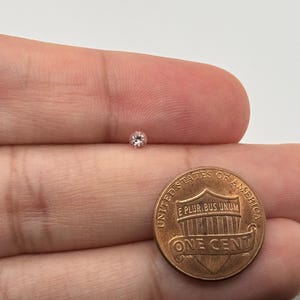 May include: A small, round, light pink diamond is shown next to a U.S. one-cent coin. The diamond is set against a neutral background, highlighting its color and shape. The coin provides a size reference for the gemstone.