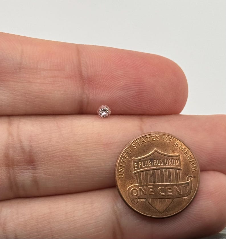 May include: A small, round, light pink diamond is shown next to a U.S. one-cent coin. The diamond is set against a neutral background, highlighting its color and shape. The coin provides a size reference for the gemstone.
