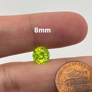 May include: A bright green, faceted gemstone, measuring 8mm in diameter, is held in a hand next to a US penny for size comparison.