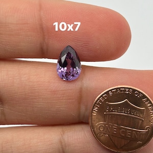 May include: A purple pear-shaped gemstone, approximately 10mm x 7mm, held in a hand for size comparison next to a US penny.