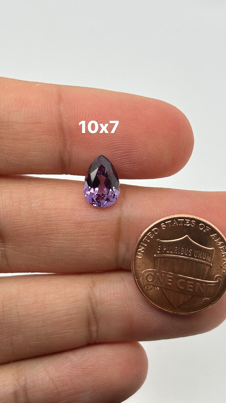 May include: A purple pear-shaped gemstone, approximately 10mm x 7mm, held in a hand for size comparison next to a US penny.