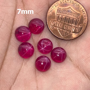 May include: Six 7mm star ruby cabochons, a type of gemstone, are displayed on a hand. The rubies are a deep red color with white star-like inclusions. A US penny is included for size comparison.