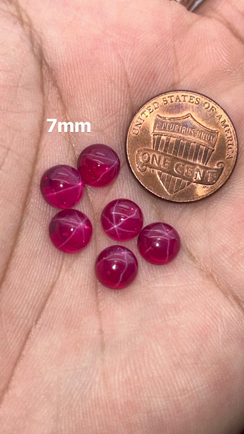 May include: Six 7mm star ruby cabochons, a type of gemstone, are displayed on a hand. The rubies are a deep red color with white star-like inclusions. A US penny is included for size comparison.