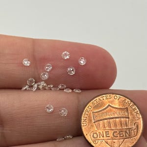 May include: A collection of small, round, clear diamonds scattered on a surface, next to a United States one-cent coin. The diamonds appear to be rose cut, with multiple facets. The coin is copper-colored and shows the words "United States of America."