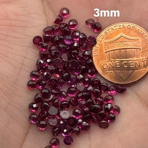 May include: A collection of small, faceted, deep red gemstones, approximately 3mm in size, are scattered across a surface. A US penny is included for size comparison. The gemstones have a brilliant cut, reflecting light.