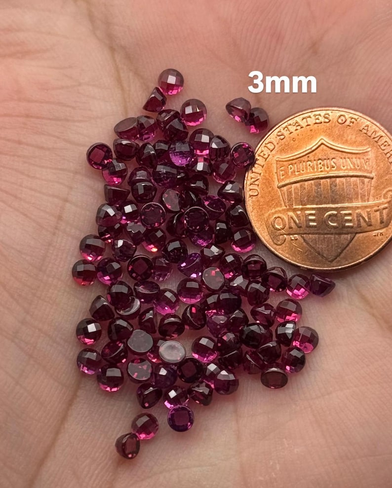 May include: A collection of small, faceted, deep red gemstones, approximately 3mm in size, are scattered across a surface. A US penny is included for size comparison. The gemstones have a brilliant cut, reflecting light.