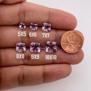 May include: A comparison of different sizes of cushion-cut amethyst gemstones, ranging from 5x5mm to 10x10mm. The gemstones are displayed on a hand, with a US penny for scale.