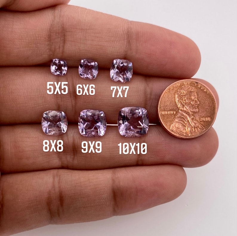 May include: A comparison of different sizes of cushion-cut amethyst gemstones, ranging from 5x5mm to 10x10mm. The gemstones are displayed on a hand, with a US penny for scale.