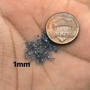 May include: A collection of small, gray gemstones, approximately 1mm in size, are displayed next to a United States one-cent coin. The gemstones are scattered, and the coin provides a size reference. The text "1mm" is visible.