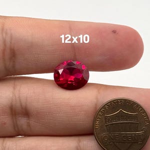 Op de afbeelding: Een gefacetteerde, ovaalvormige rode edelsteen met een levendige karmozijnrode tint. De steen is geplaatst naast een Amerikaanse munt van één cent voor schaal, met de tekst 12x10 boven de edelsteen, wat de afmetingen in millimeters aangeeft.
