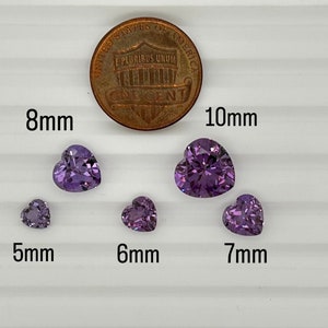 K&ouml;nnte beinhalten: F&uuml;nf violette herzf&ouml;rmige Edelsteine ​​in verschiedenen Gr&ouml;&szlig;en von 5 mm bis 10 mm werden auf einer wei&szlig;en Oberfl&auml;che neben einem 1-Cent-St&uuml;ck zum Gr&ouml;&szlig;envergleich angezeigt.