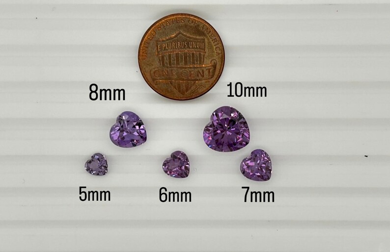 K&ouml;nnte beinhalten: F&uuml;nf violette herzf&ouml;rmige Edelsteine ​​in verschiedenen Gr&ouml;&szlig;en von 5 mm bis 10 mm werden auf einer wei&szlig;en Oberfl&auml;che neben einem 1-Cent-St&uuml;ck zum Gr&ouml;&szlig;envergleich angezeigt.