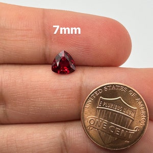May include: A 7mm red triangular gemstone held in a hand next to a US penny.