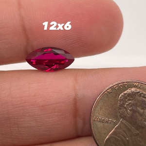 May include: A deep red marquise-shaped gemstone, measuring 12x6, is held in a hand for size comparison. A US penny is also shown for scale.