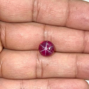 May include: A round, polished, star ruby gemstone. The ruby is a deep, vibrant red with a six-rayed star pattern across its surface. The gemstone is held in a hand, showcasing its color and clarity.