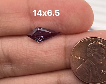 Può includere: Una pietra preziosa viola con un taglio a forma di aquilone sfaccettato. La pietra preziosa è tenuta in una mano accanto a un centesimo americano per il confronto delle dimensioni. Il testo "14x6.5" è visibile nell'immagine.