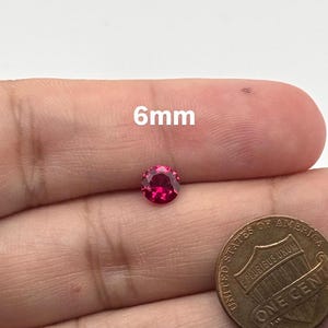 May include: A round, faceted, vibrant pinkish-red gemstone measuring 6mm in diameter, positioned next to a United States one-cent copper coin for scale. The gemstone displays a brilliant cut with multiple reflective surfaces.