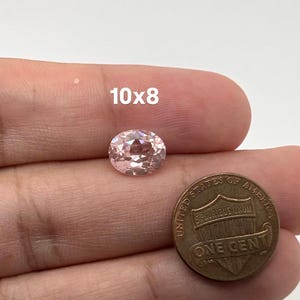 May include: An oval-cut, light pink gemstone with a faceted surface. The stone is positioned next to a United States one-cent coin for scale, with the text 10x8 printed above the gemstone, indicating its dimensions in millimeters.