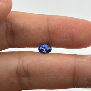 May include: A deep blue oval-shaped gemstone, likely a sapphire, is held between two fingers.