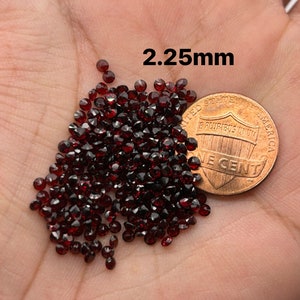 May include: A pile of small, dark red gemstones, approximately 2.25 mm in size, next to a US penny for scale.