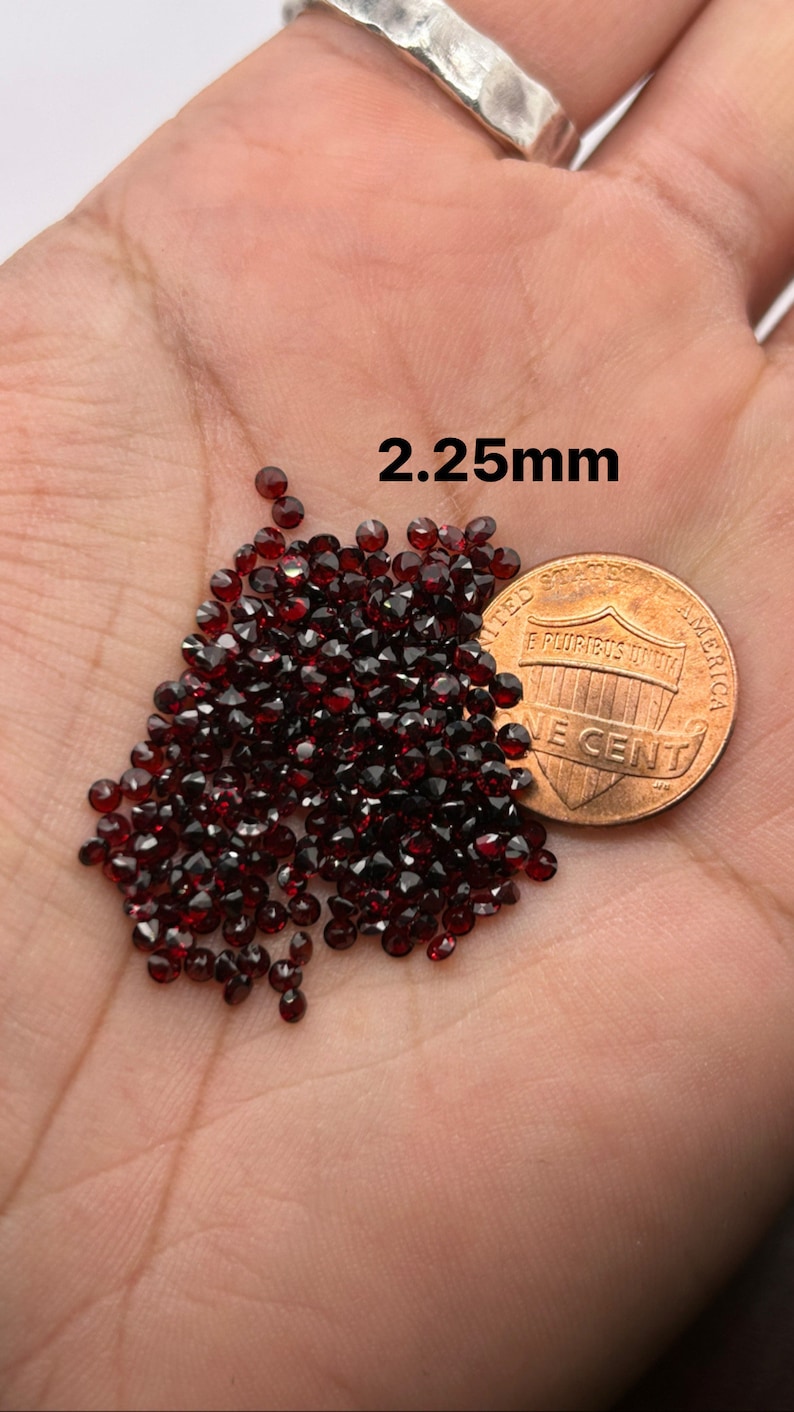 May include: A pile of small, dark red gemstones, approximately 2.25 mm in size, next to a US penny for scale.