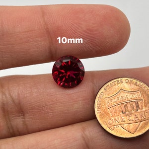 May include: A faceted, round, deep red gemstone, approximately 10mm in diameter, is held between fingers. A U.S. one-cent coin is next to the gemstone for size comparison. The text "10mm" is above the gemstone.