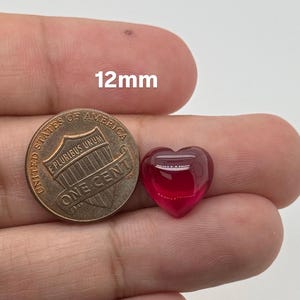 May include: A small, smooth, translucent red heart-shaped gemstone next to a United States one-cent coin. The text 12mm is visible above the coin, indicating the size of the heart-shaped object in millimeters for scale and reference.