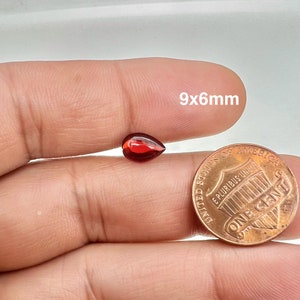 May include: A red pear-shaped gemstone measuring 9x6mm. The gemstone is held in a hand for size comparison next to a US penny.