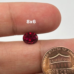 May include: A deep red oval gemstone, approximately 8mm by 6mm, is held in a hand next to a US penny for size comparison.