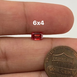 May include: A red gemstone with a rectangular emerald cut, measuring 6x4 millimeters. The gemstone is being held in a hand, next to a US penny for size comparison.