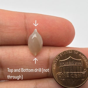 May include: A light gray, teardrop-shaped stone with a drilled hole at the top and bottom. The stone is being held next to a US penny for size comparison.