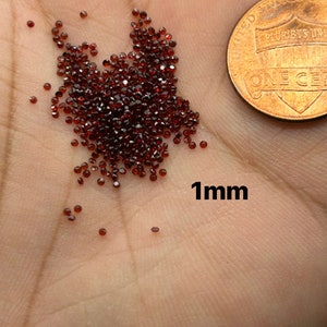 May include: A pile of small, dark red, faceted gemstones, approximately 1 mm in size, on a person's hand. A US penny is in the background for scale.