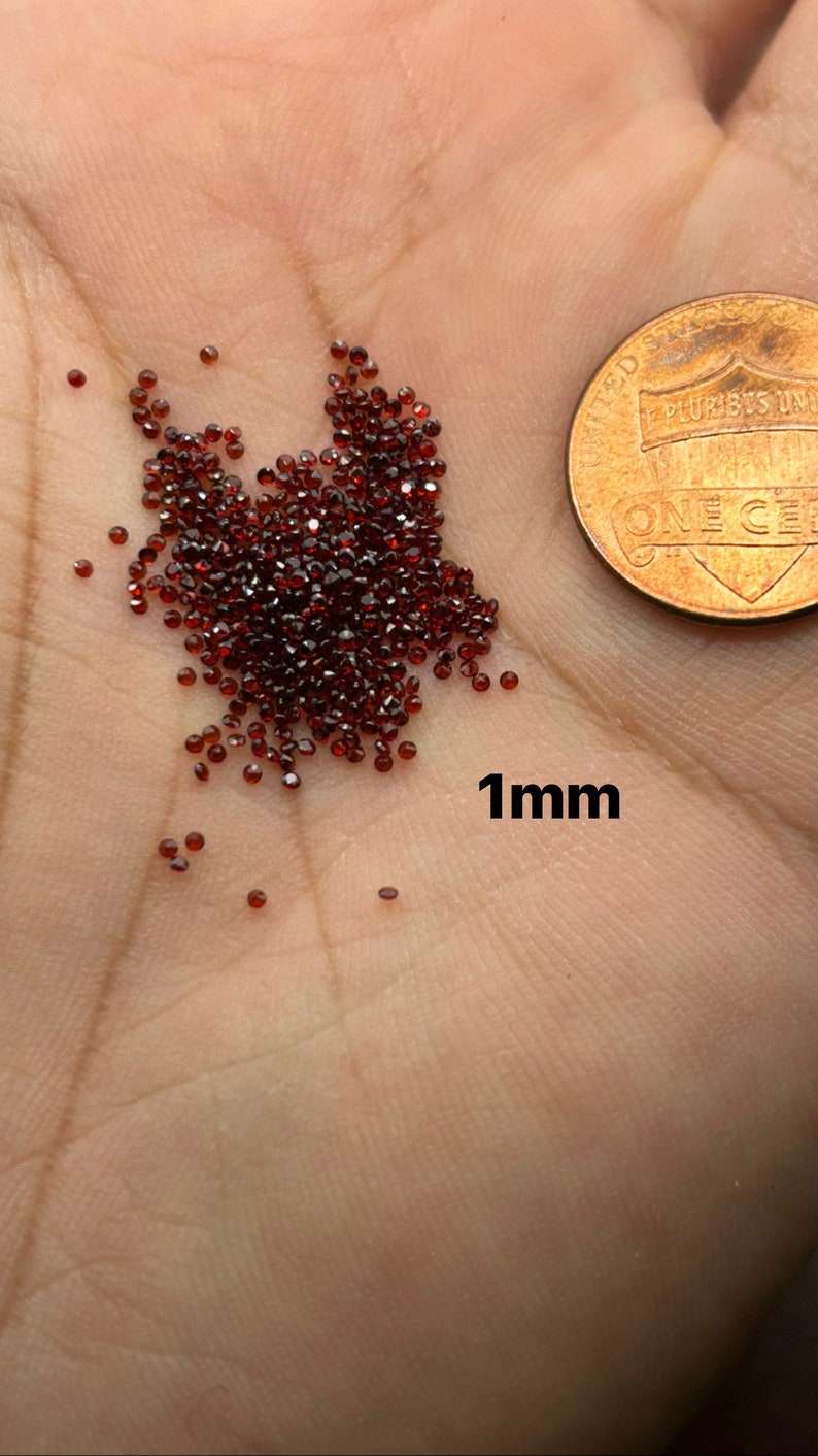 May include: A pile of small, dark red, faceted gemstones, approximately 1 mm in size, on a person's hand. A US penny is in the background for scale.