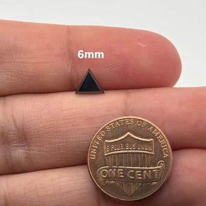 Może przedstawiać: Czarny, trójkątny kolczyk o wymiarach 6 mm. Kolczyk jest pokazany na palcu osoby obok amerykańskiego centa.