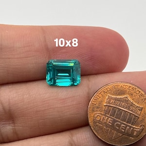 Puede incluir: Una gema de esmeralda verde vibrante con un corte esmeralda rectangular. La gema mide aproximadamente 10x8 milímetros y se muestra junto a un centavo de dólar estadounidense para la escala.