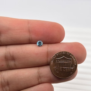 K&ouml;nnte beinhalten: Ein kleiner, hellblauer Edelstein wird in einer Hand neben einem US-Penny gehalten. Der Penny ist eine Kupferm&uuml;nze mit den Worten "United States of America" und "One Cent" darauf.