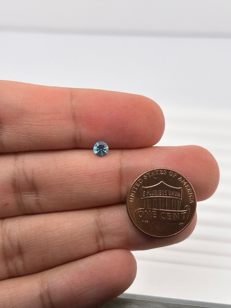 K&ouml;nnte beinhalten: Ein kleiner, hellblauer Edelstein wird in einer Hand neben einem US-Penny gehalten. Der Penny ist eine Kupferm&uuml;nze mit den Worten "United States of America" und "One Cent" darauf.