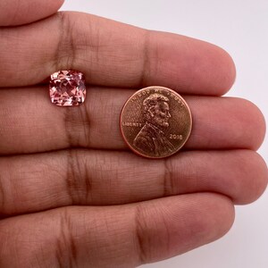 May include: A pink gemstone, approximately the size of a US penny, is held in a hand. The gemstone is a square cushion cut with facets.