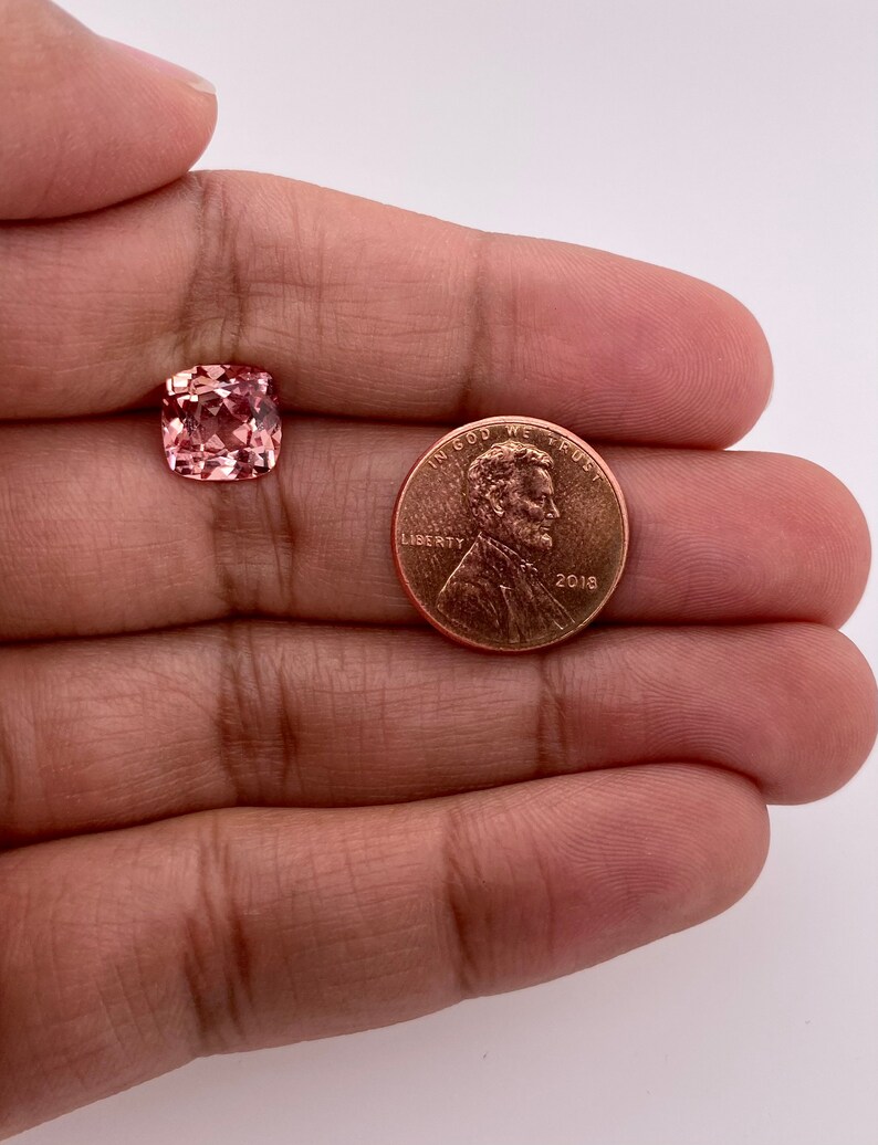 May include: A pink gemstone, approximately the size of a US penny, is held in a hand. The gemstone is a square cushion cut with facets.