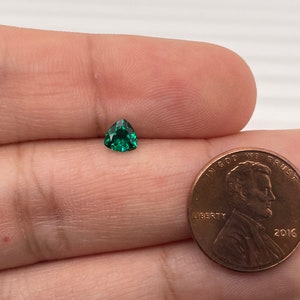 May include: A small, triangular, green gemstone, approximately the size of a US penny.