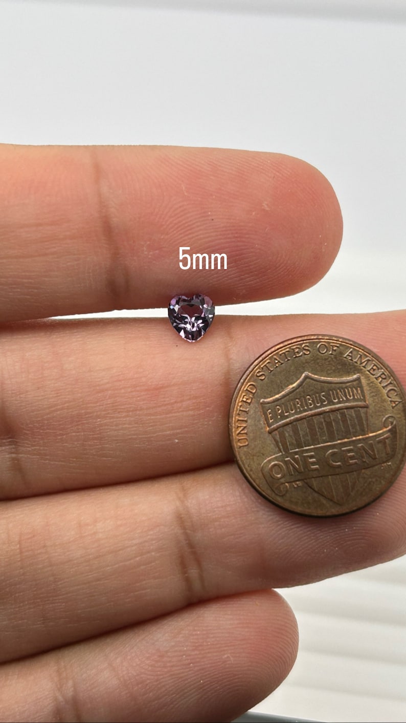 K&ouml;nnte beinhalten: Ein lilafarbener herzf&ouml;rmiger Edelstein mit einem Durchmesser von 5 mm. Der Edelstein wird von einer Hand gehalten, mit einem US-Penny als Gr&ouml;&szlig;envergleich.