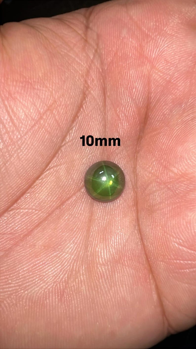 May include: A round, green gemstone, approximately 10mm in diameter, rests in an open palm. The gem has a star-like pattern within. The text "10mm" is displayed above the gem.