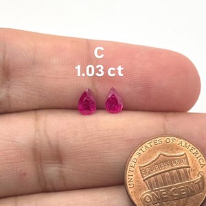 Puede incluir: Dos piedras preciosas facetadas en forma de pera, de un vibrante color rojo rosado, etiquetadas como C con un peso total de 1.03 ct. Una moneda de un centavo de los Estados Unidos se coloca junto a las piedras para dar una escala visual.