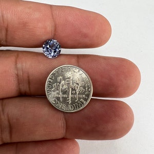 Peut inclure: Une pierre précieuse ronde, facettée et de couleur violet clair, placée à côté d'une pièce de dix cents des États-Unis pour l'échelle. La pierre présente une taille brillante claire avec de multiples facettes reflétant la lumière.