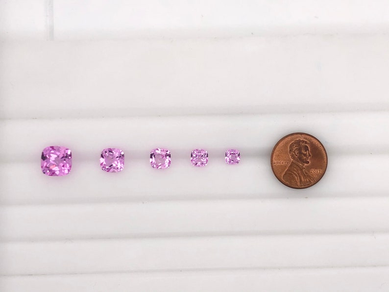 May include: Five pink square-shaped gemstones of varying sizes, arranged in a row from largest to smallest. A penny is included for scale.