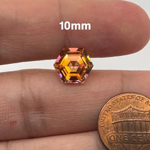 May include: A 10mm hexagonal cut gemstone with a mix of orange, yellow, and pink hues. The gemstone is displayed on a finger for size comparison.