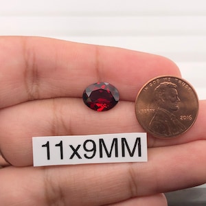 May include: A deep red oval gemstone measuring 11x9mm. The gemstone is being held in a hand next to a US penny for size comparison.